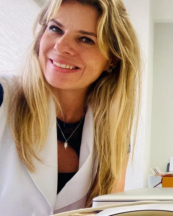 Simone Tavolaro Luisada sorrindo, sentada à mesa de atendimento em seu consultório